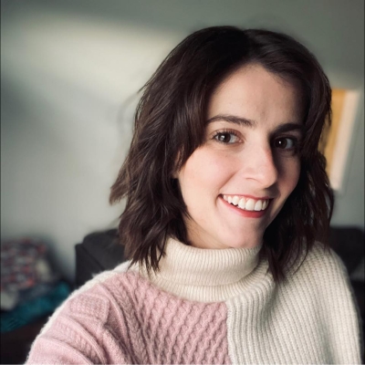 Woman with brown hair wearing a pink and white sweater in front of a grey background.